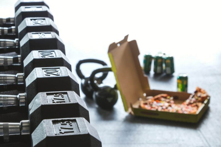Una caja de pizza desenfocada en el suelo del gimnasio con seis latas al lado.